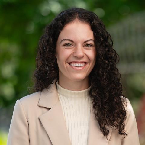 This is an image of Angelica, who is standing outdoors in a park. She has long dark, curly hair and is wearing a mock neck beige top with a blazer. The background includes dark green green foliage.