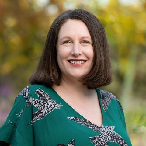 This is an image of Nina, a individual smiling in a park environment. She has shoulder length dark hair and is wearing a green blouse with a bird print. The background includes vibrant green and gold foliage.