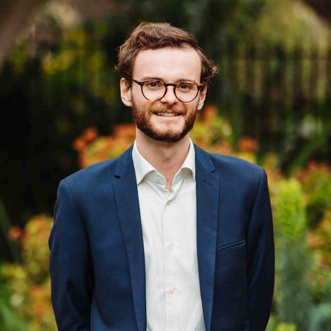 A photo of Mat who has short brown hair, glasses, and a beard is standing outdoors, smiling at the camera. He is wearing a white shirt and a dark blue blazer. The background is blurred greenery with hints of orange flowers.