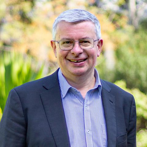 This is an image of Matthew, a smiling individual standing in a park. He has short, silver hair, is wearing a pair of glasses and a checkered collared shirt with a blazer. The background includes vibrant green foliage.