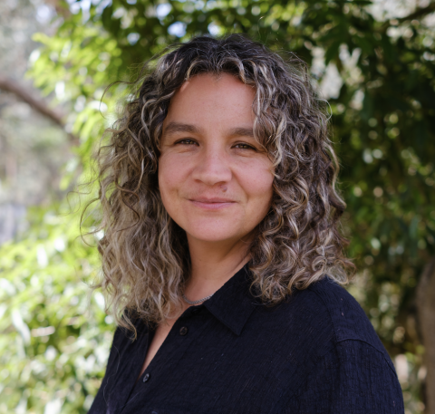This is an image of Vanessa, a smiling individual with curly brown-blonde hair. Vanessa is standing outside, in front of green trees on a sunny day.