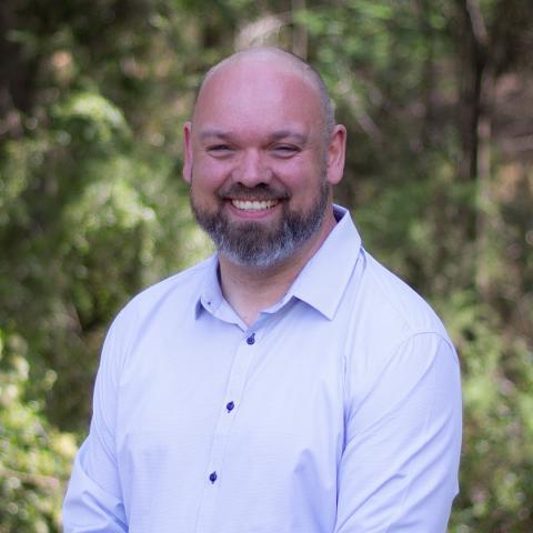 This is an image of Andrew, who smiles brightly at the camera in front of a blurred bushland environment. He has a shaved head, and a short, salt and pepper beard. He wears a white collared shirt with dark buttons.