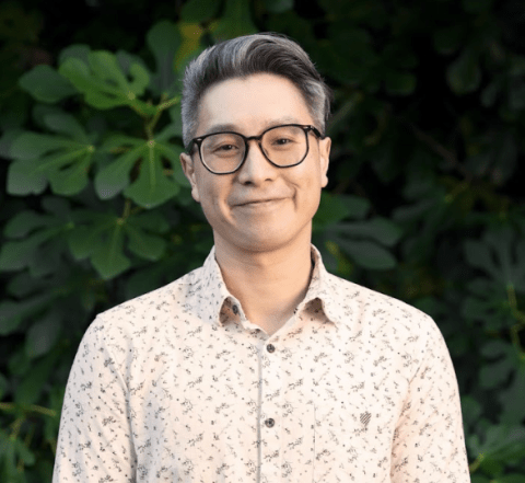 This is an image of Hsiang, a smiling individual with glasses and black-grey hair. Hsiang is wearing a collared shirt and standing outside, in front of dark green shrubbery.