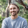 This is an image of Mitch, a smiling individual. Mitch has brown hair and is wearing a grey long sleeve shirt. He is standing outside in front of green trees on a sunny day. 