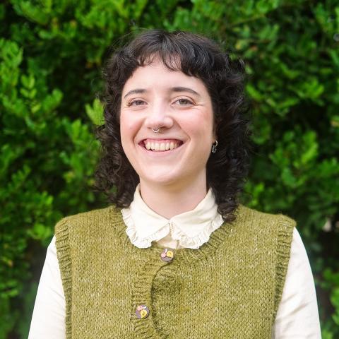 This is an image of Ebony, who is smiling at the camera in front of a lush green hedge. She has dark curly hair that sits on her shoulder, with a short fringe that frames her forehead. She wears a light coloured blouse with an olive, knitted vest and has a silver septum piercing and a small silver hoop in her left ear.