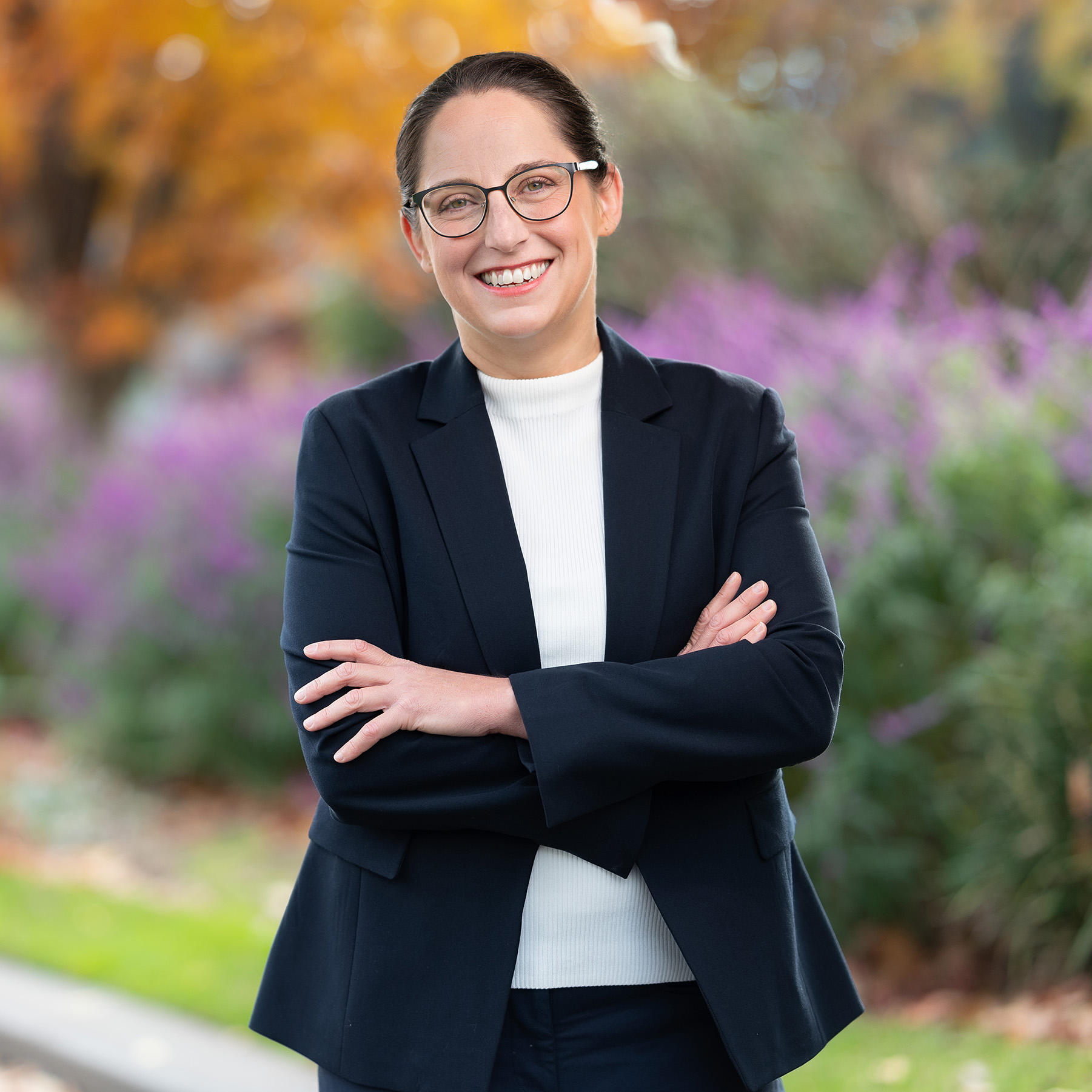 Melanie is a smiling woman with glasses and dark hair pulled back, wearing a navy blazer and white top, standing outdoors with arms crossed in front of a background of purple flowers and autumn-coloured trees.