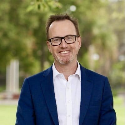 David is wearng a white shirt and blue jacket, glasses and smiling at the camera