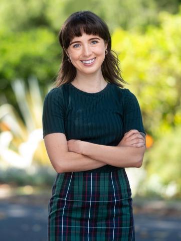 A smiling woman stands confidently outdoors with arms crossed. She wears a dark green ribbed top paired with a green plaid skirt. Her straight brown hair frames her face, and a lush, sunlit garden forms the background.