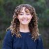 This is an image of Sophie, a brightly smiling individual standing in a bushland environment. She has brown curly hair with a fringe that sits just below her shoulders and wears a navy shirt. The background is blurred dark green, creating a calm and pleasant setting.
