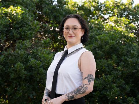 Profile picture of Greens candidate for South-West Coast, Silver Rose. Silver is standing in front of bushes and is wearing a sleaveless white shirt with a thin black tie. Silver is standing slightly to the side while looking at the camera