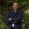 This is an image of Binoy, a smiling individual with a dark blue coat, a light blue collared shirt and denim jeans. He is standing outdoors, in front of dark green trees.