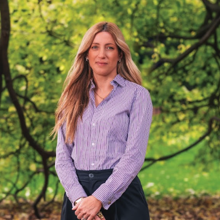 This is an image of Hayley, a smiling individual with long, blonde hair. Hayley is wearing a purple button down collared shirt and dark trousers, and is standing outside, in front of sun-lit green trees. 