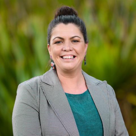 This is an image of Alysia, who is smiling brightly at the camera in front of a lush, green, outdoor environment. Alysia has dark brown hair tied up in a bun and wears a grey blazer, blue textured blouse and brightly coloured earrings that have a textured paint brush design.