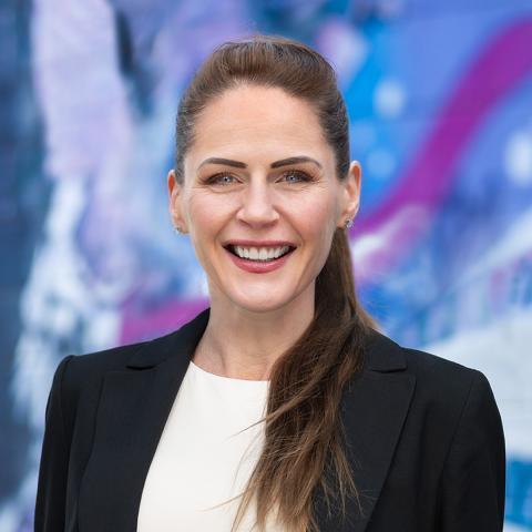 This is an image of Sonya, who smiles warmly at the camera. She's standing outdoors in front of a wall with a blue & purple mural painted on it. Sonya has long, brown hair tied into a ponytail that falls on her left shoulder. She wears a white blouse and black blazer with stud earrings.