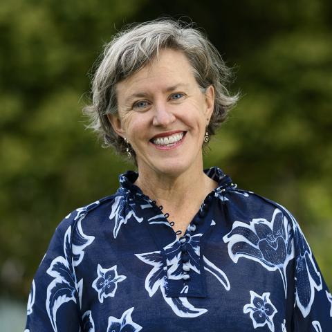Profile picture of Cate Sinclair, lead Greens candidate for Northern Victoria upper house region. Cate is smiling while wearing a dark blue top with flowers that are outlined in white and is standing in front of blurred out bushes