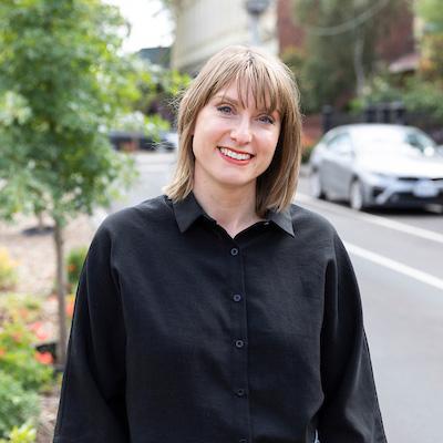 This is an image of Rachel, she's smiling warmly at the camera. She has shoulder-length, light brown hair with bangs and stands in an inner city streetscape.