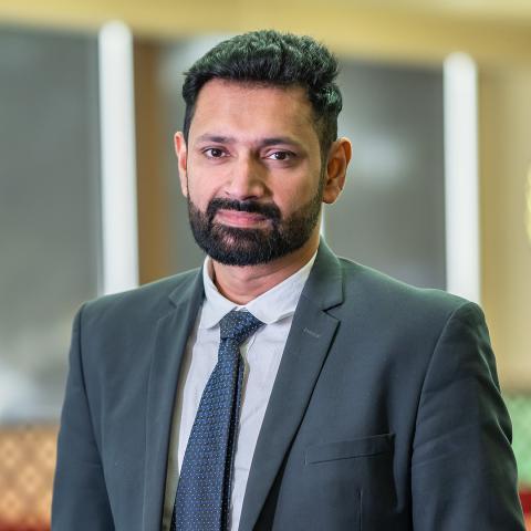 This is an image of Tushar, a smiling person in a charcoal blazer, white collared shirt and dark tie who is standing in an office environment. He has short, dark hair and a neatly trimmed beard.
