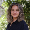 This is an image of Vanessa, a smiling individual with curly brown-blonde hair. Vanessa is standing outside, in front of green trees on a sunny day.