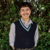 This is an image of Kai, a smiling individual standing outside, in front of dark green trees. Kai is wearing a collared white and blue striped shirt with a dark blue vest pullover on top.