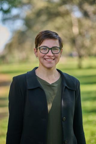A woman with short brown hair and glasses, wearing a dark green shirt and blazer standing in a park smiling