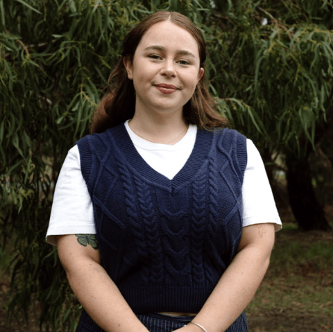 This is an image of Zoe, a smiling individual with long brown hair, a white t-shirt and a blue knitted vest. She is standing outdoors, in front of dark green bushes.