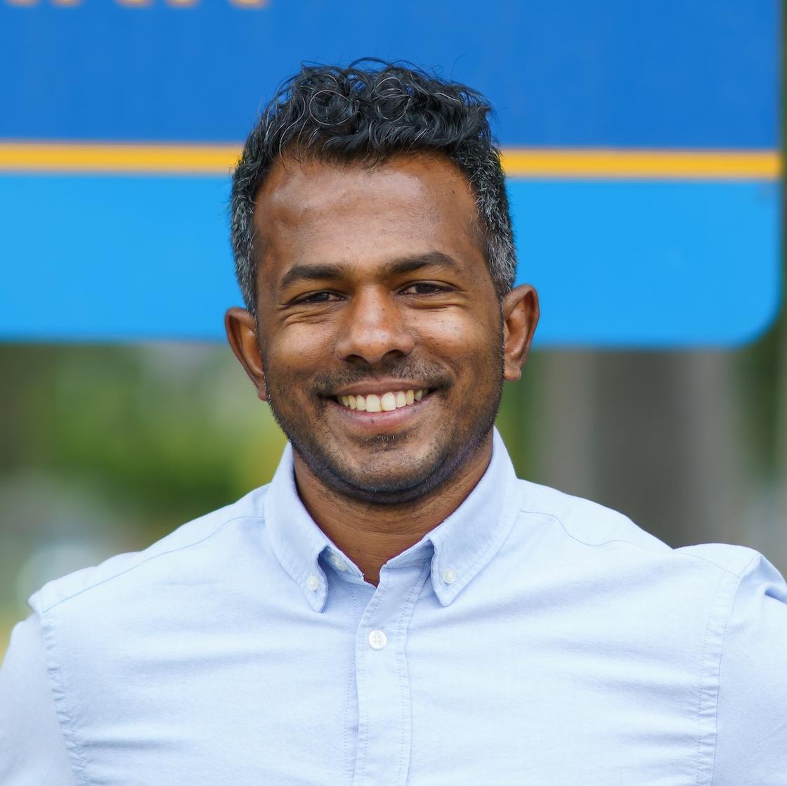Sujan is wearing a light blue shirt, smiling at the camera and is situated outside 