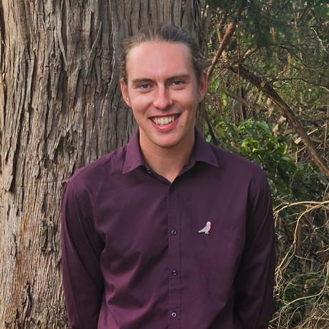 This is an image of Zave, who is smiling at the camera in front of a large tree and bushland. He has long, brown hair that is tied up and is wearing a purple collared shirt that features a small bird where a pocket would be.