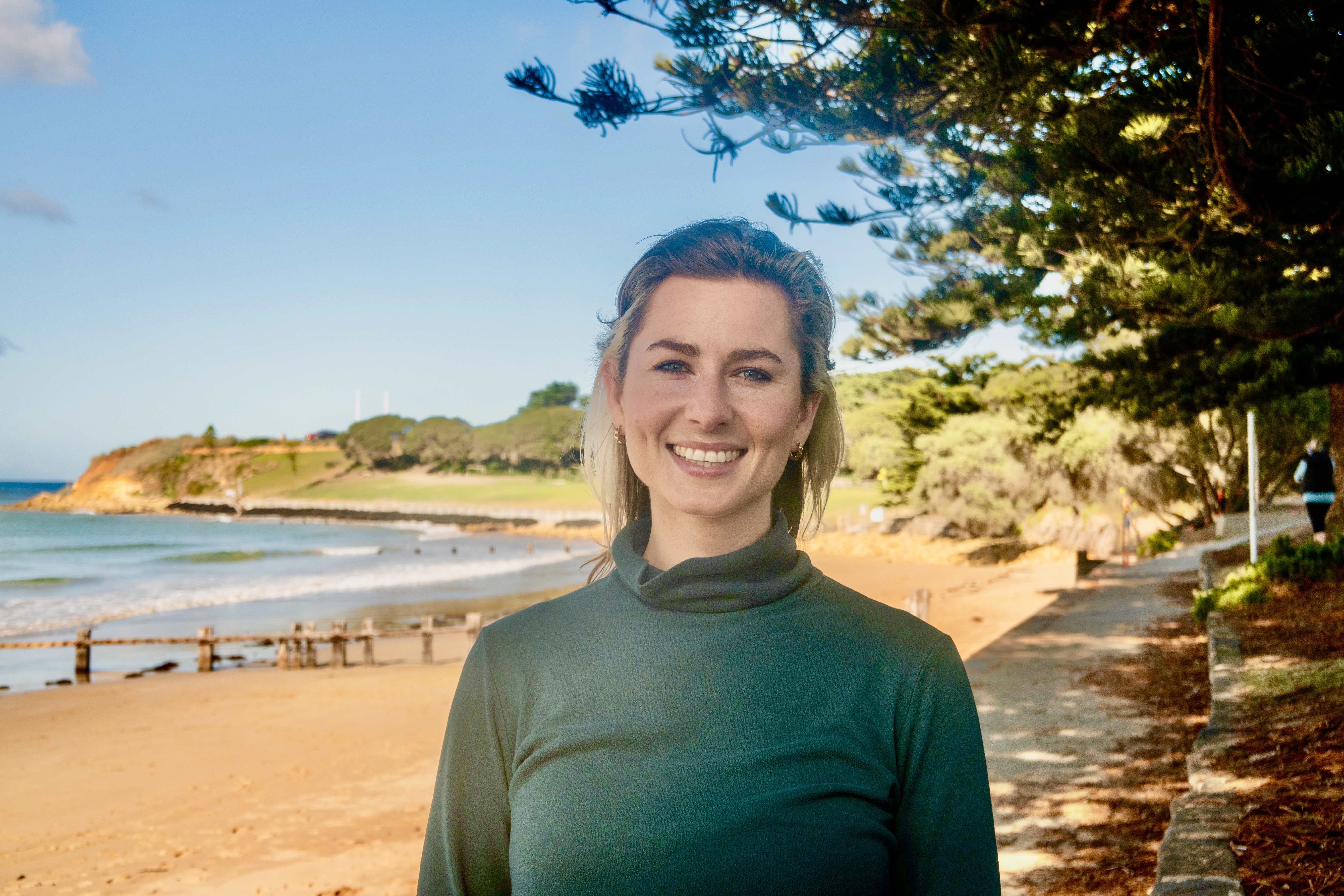 Phoebe stands in front of a beach, smiling warmly. She wears a dark green turtleneck, and has shoulder length dark blonde hair.