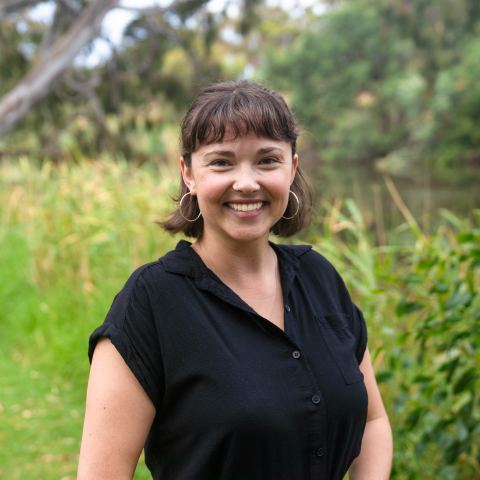 This is an image of Emilie, a smiling individual standing outdoors in a lush natural environment. She has short dark hair with bangs, is wearing a black collared shirt, and accessorized with large hoop earrings. The background includes vibrant green foliage, tall grasses, and a serene pond or wetland area, creating a calm and pleasant setting.