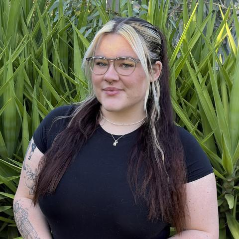 This is an image of Grace, who smiles at the camera in front of some bright greenery. Her hair is long, dark brown with blonde highlights that frame her bespectacled face. She wears a black tshirt so you can see some tattoos along one upper arm, and two small pendants around her neck.