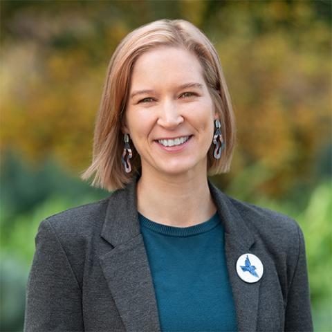 Profile picture of Nina Laitala, Greens candidate for Bundoora. Nina is wearing a grey jacket with a blue suit and is wearing a white badge with a blue bird on it. She is standing in front blurred out parklands.