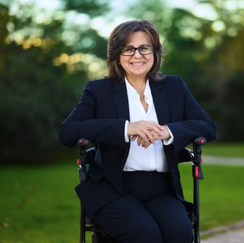This is an image of Georgia, a smiling individual with dark hair, glasses and wearing a white blouse with a black blazer. Georgia is sitting on a seat walker, outside in the greenery. The background is full of dark and light green trees and sunlight.