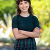 A smiling woman stands confidently outdoors with arms crossed. She wears a dark green ribbed top paired with a green plaid skirt. Her straight brown hair frames her face, and a lush, sunlit garden forms the background.