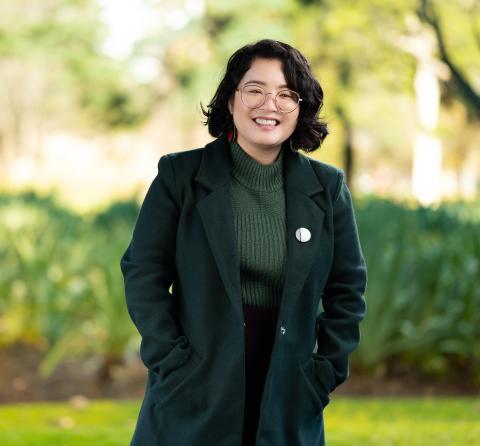 A woman with shoulder-length brown hair, glasses, wearing a green jacket and smiling in a green park.