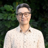 This is an image of Hsiang, a smiling individual with glasses and black-grey hair. Hsiang is wearing a collared shirt and standing outside, in front of dark green shrubbery.