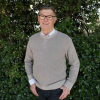 This is an image of Dave, a smiling individual. Dave is wearing glasses, a pullover and a collared shirt. He is standing in front of dark green trees on a sunny day.