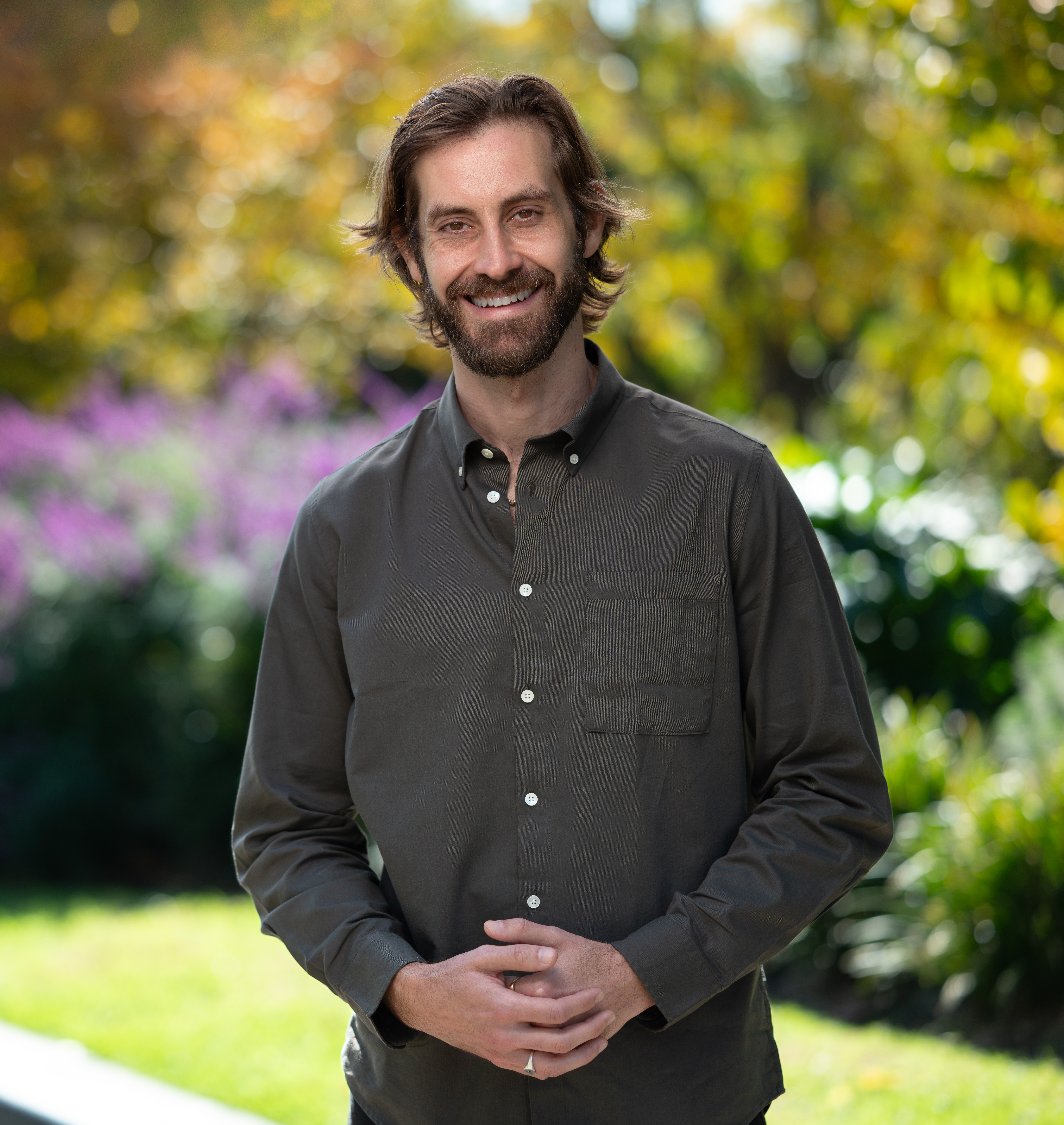A man with dark brown hair, a brown beard, wearing a dark green shirt smiling in front of a leafy background.