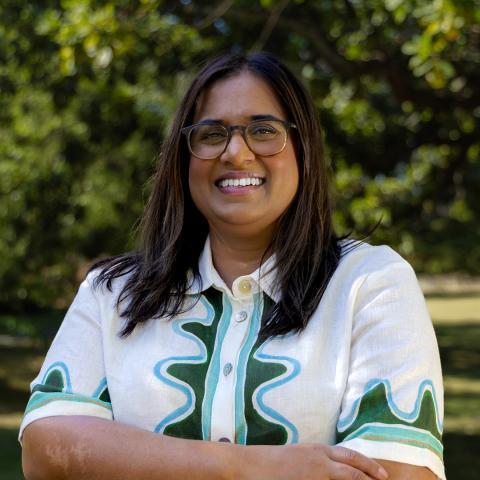 This is an image of Kylie, a brightly smiling individual standing outdoors. She has dark hair that sits below her shoulders, is wearing a collared short sleeve shirt that is mostly white with a green pattern at the hems. Glasses frame her face.