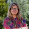 This is an image of Veronika, who smiles warmly at the camera. She is standing outdoors in an outdoor environment, blurred trees fill the background. She has shoulder-length, blonde wavy hair, and wears a floral printed shirt of pink and purple. Black glasses frame her face.