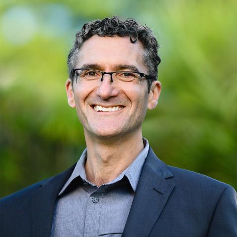 This is an image of Campbell, a smiling individual standing outdoors in a lush natural environment. He has short, salt & pepper curls and is wearing a pair of glasses, a petrol blue collared shirt, and navy jacket. The background features a vibrant green gradient that is out of focus and helps to make the subject pop in the shot.