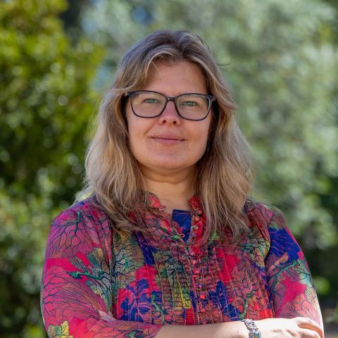 This is an image of Veronika, who smiles warmly at the camera. She is standing outdoors in an outdoor environment, blurred trees fill the background. She has shoulder-length, blonde wavy hair, and wears a floral printed shirt of pink and purple. Black glasses frame her face.