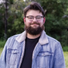 This is an image of Gavin, a smiling individual with dark hair, a beard and moustache. Gavin is standing outside in front of dark green trees and is smiling kindly. 
