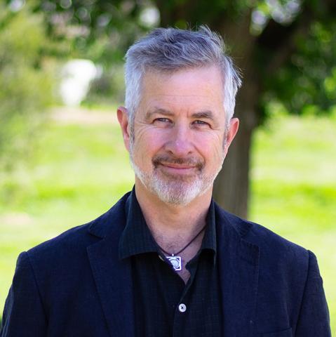 This is an image of Tim who smiles at the camera in front of a blurred park environment. He has short salt & pepper hair and stubble, and wears a dark collared shirt. A silver pendant with a black rope chain is around his neck.