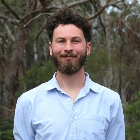 This image depicts Jesse who is stood smiling, outdoors in a coastal bushland environment. He has short, dark, wavy hair and a neat diamond beard that features some lighter areas. Jesse is wearing a light blue collared shirt, and has a stud earring in his left lobe. The background includes tall trees with their brown branches sprawling and green shrubbery, with a small amount of blue sky peeking through.