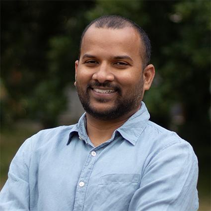 This is an image of Gufran, a smiling individual standing outdoors. He has a mostly shaved head, with some dark hair and is wearing a light blue collared shirt.