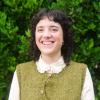 This is an image of Ebony, who is smiling at the camera in front of a lush green hedge. She has dark curly hair that sits on her shoulder, with a short fringe that frames her forehead. She wears a light coloured blouse with an olive, knitted vest and has a silver septum piercing and a small silver hoop in her left ear.