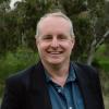 This is an image of Ben, who appears to be in a bushy, outdoor environment. He has short, grey hair and is smiling brightly at the camera whilst wearing a black jacket and a dark bluey-green shirt.