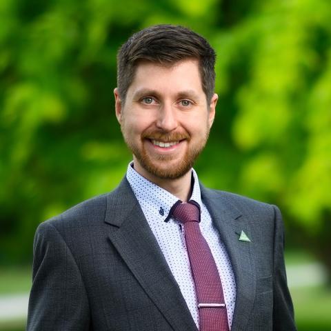 This is an image of Alex, who smiles warmly at the camera in front of a bright green outdoor environment. Alex wears a dark grey jacket with a collared shirt and burgundy tie. A Greens badge is pinned to his lapel. He has dark hair and a short reddish beard..