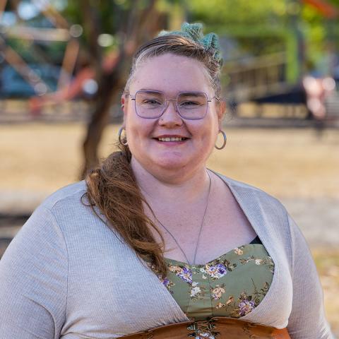 This is an image of Theresa, who smiles at the camera in a park environment that is blurred behind her. Her long auburn-coloured hair is tied up in a ponytail with a teal coloured scrunchie and she wears a green floral sundress under a beige cardigan. There is a long pendant around her neck, hoops in both her ears and she wears a pair of square, purple frames.