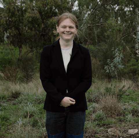 This is an image of Joyce, a smiling individual with a black jacket. Joyce is standing outside, and in the background there are green trees and shrubbery.